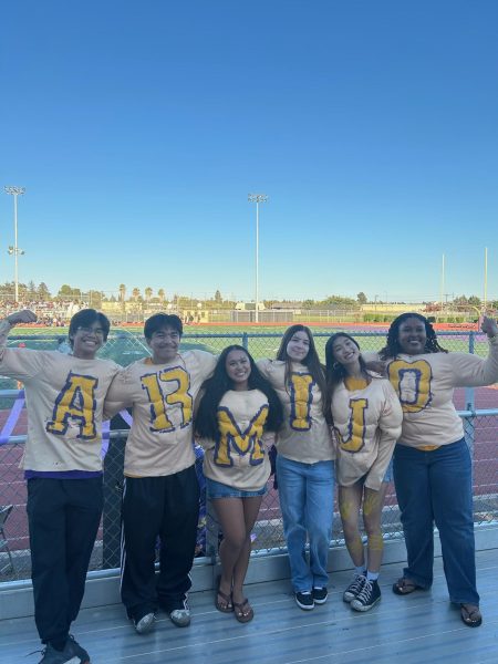 Bold Spirit on Display
The 6 Senior ASB Officers [Mark Silva], [Ethan Magno], [Mariza Supapo], [Kiera Tomko], [Ahri Aguba], and [Osaguina Sorae] arrive early to the game to help set up and boost school spirit, gathering at the fence to hype up the crowd as the sun sets behind the bleachers. Their coordinated shirts, painted by ASB officers in the past years, become a highlight of the event as students stop to take photo with them. The moment captures how school events bring students together through shared pride and energy.
